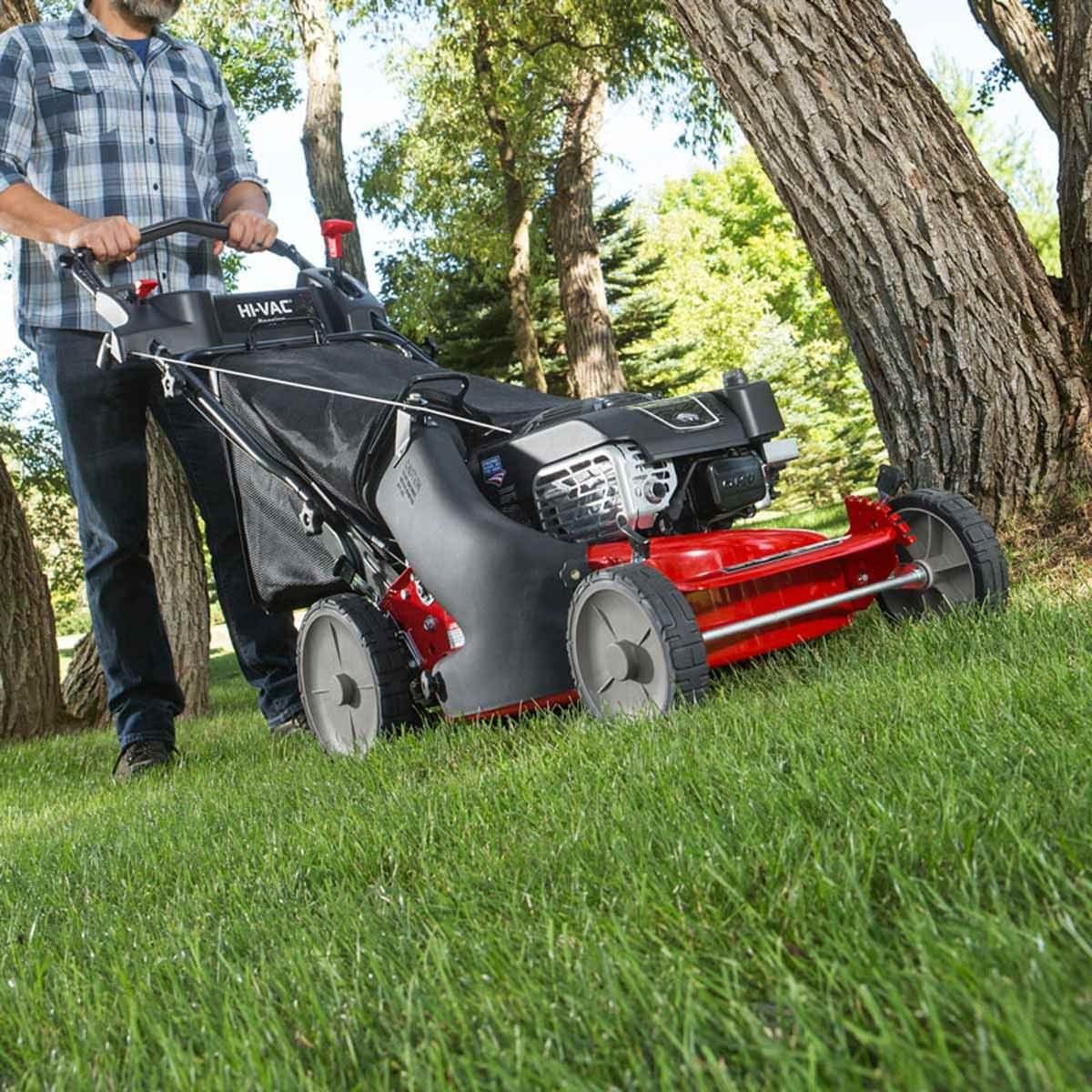 A person operates a red lawn mower, trimming grass in a green outdoor area surrounded by trees.