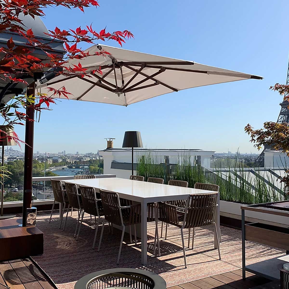 A large dining table with chairs sits under a white umbrella on a rooftop terrace, surrounded by plants and a clear blue sky, with the Eiffe