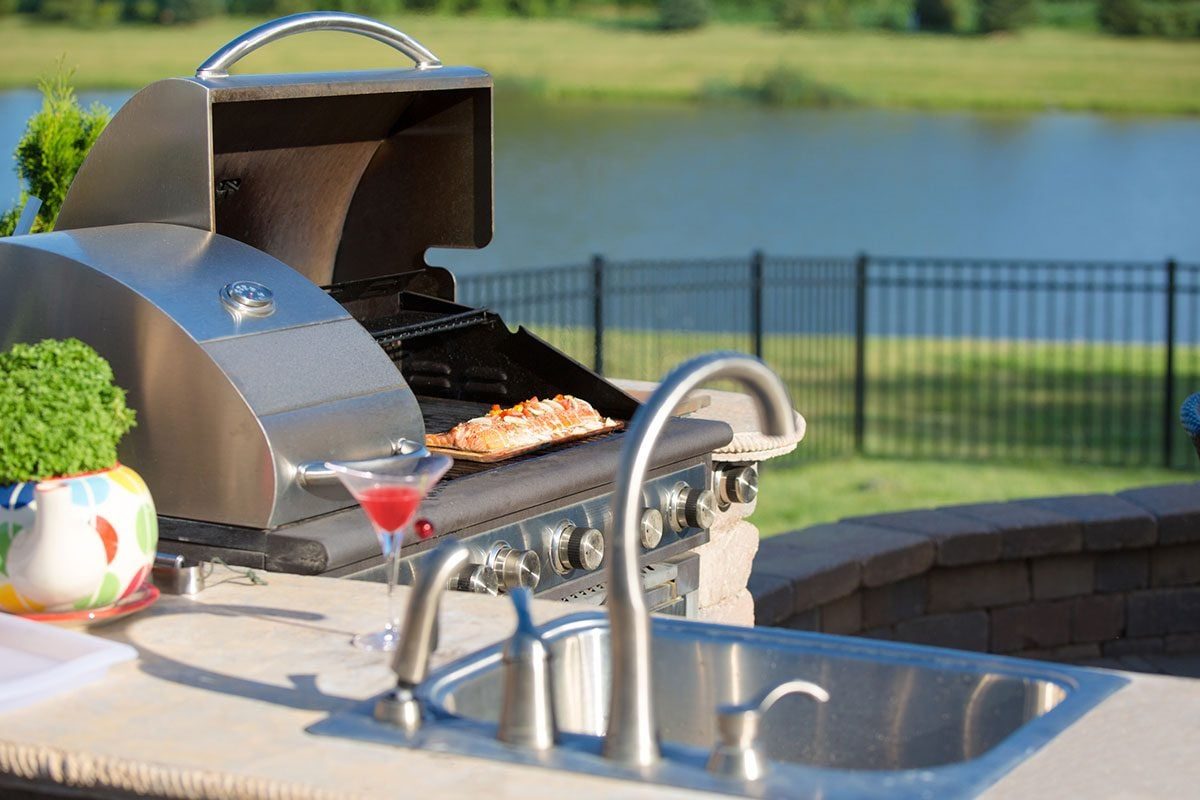 A shiny grill cooks salmon in an outdoor kitchen setting, featuring a cocktail and sink, with a grassy area and fence near a body of water.