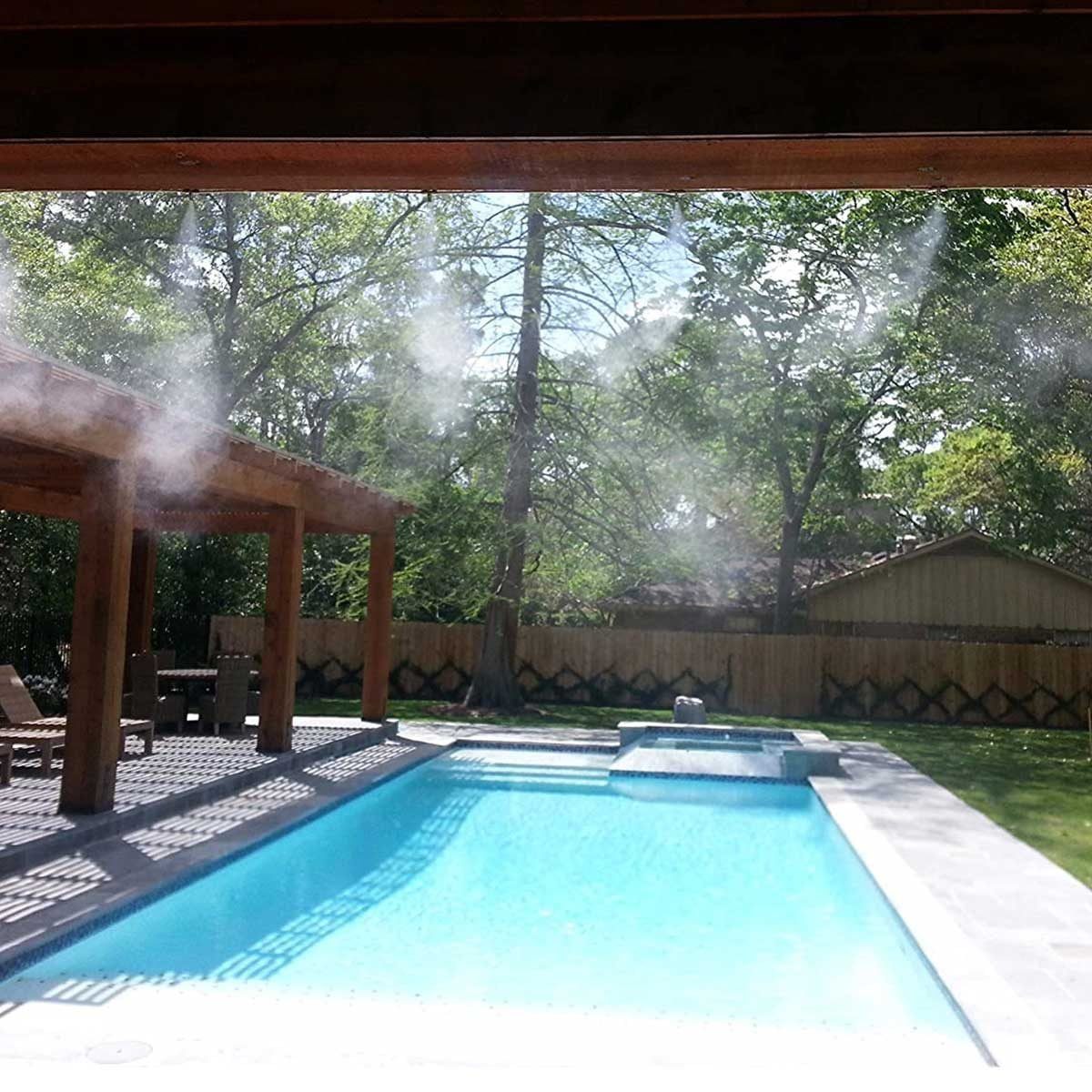 A misting system releases water vapor above a clear swimming pool, surrounded by wooden structures and a lush green landscape under a bright