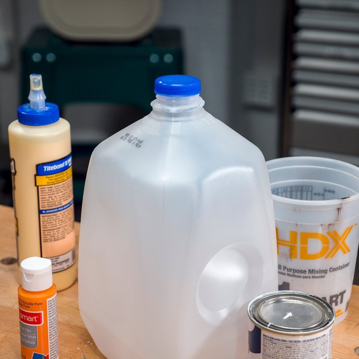 A large, translucent plastic jug with a blue cap sits on a wooden surface alongside various containers, indicating a workspace for crafting