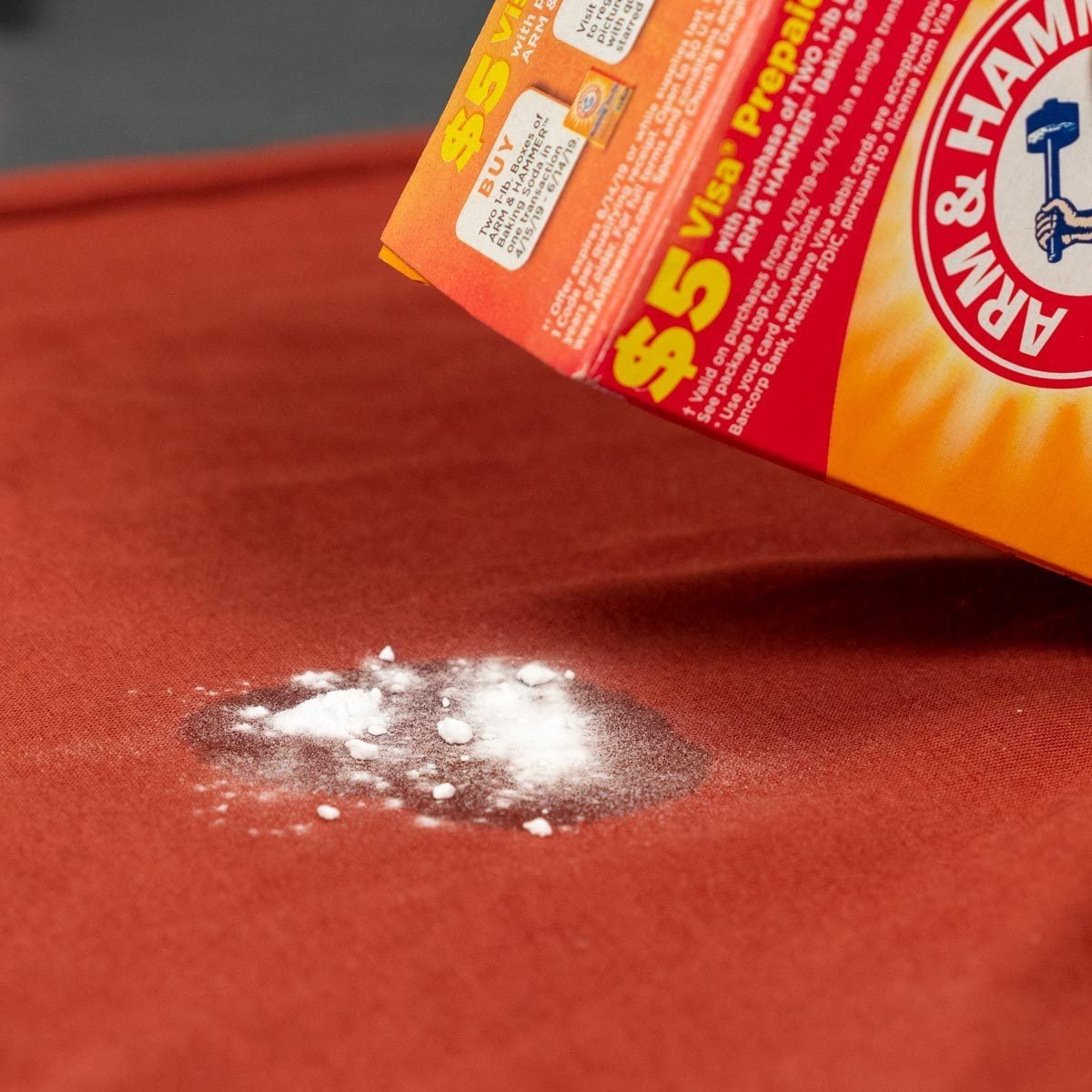 A box is pouring white powder onto a reddish-brown fabric surface, creating a small, scattered pile of powder.