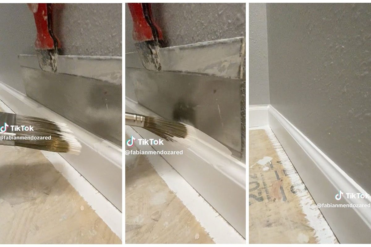 A paintbrush applies white paint along a baseboard, while a putty knife remains nearby on a bare floor, set against a gray wall.
