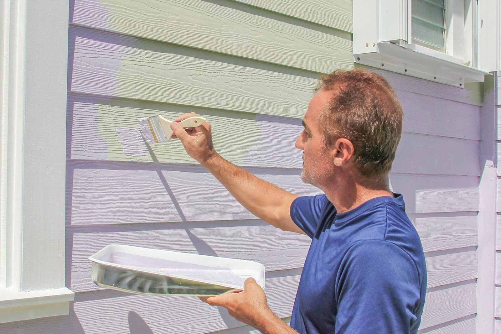 A man paints a house exterior using a small roller, while holding a tray of paint, under bright sunlight. The wall features varying shades o