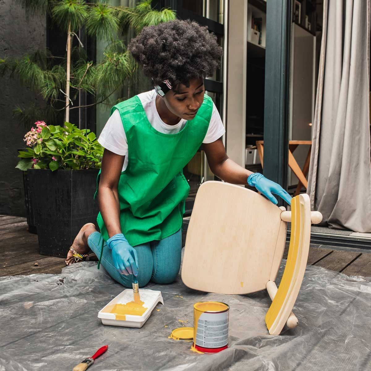 A person in a green apron paints a wooden chair with yellow paint, surrounded by a garden, on a tarp in a light-filled outdoor area.