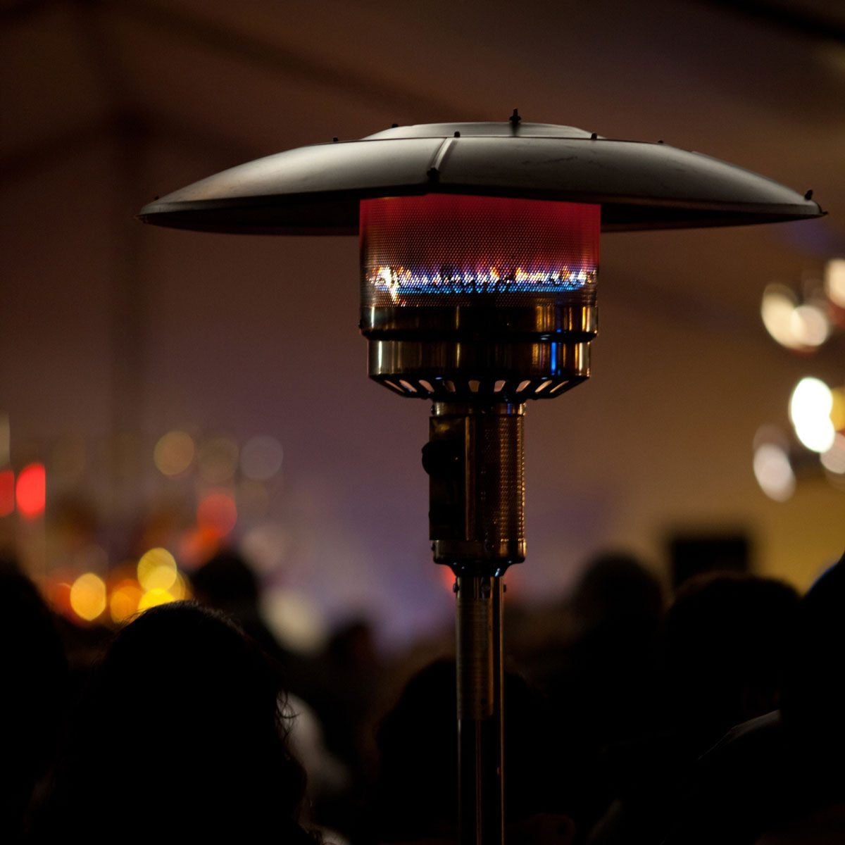 A gas patio heater radiates warm light in a dimly lit area, surrounded by blurred silhouettes of people and soft, colorful background lights