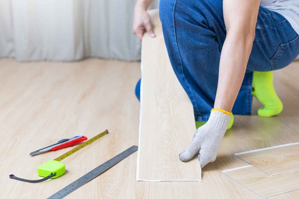 A person in gloves positions a plank of flooring on a wooden surface, surrounded by measuring tools and materials for home improvement.