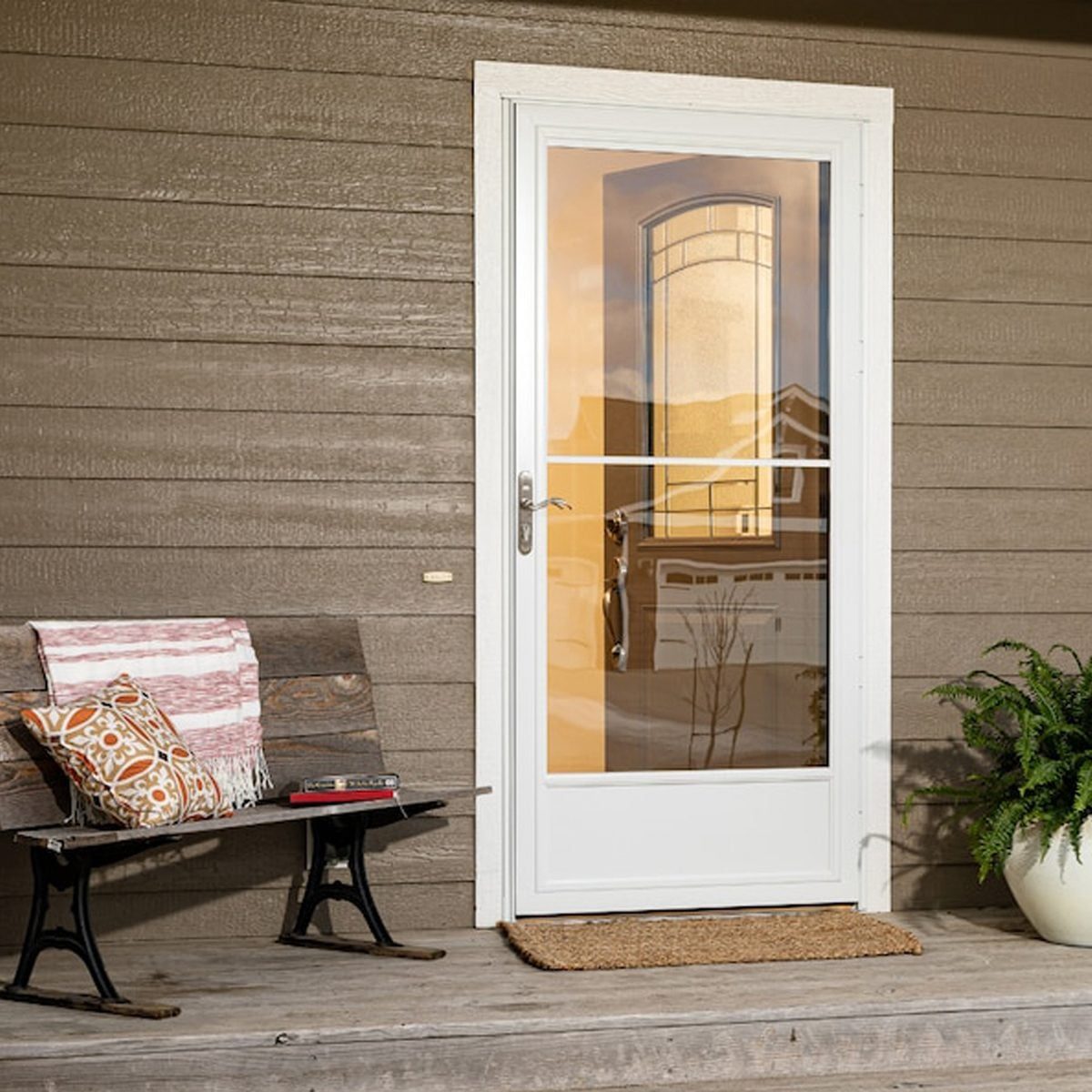 A white door stands closed, framed by wooden siding. A bench and potted plant sit nearby, while a reflection of a house is visible in the do
