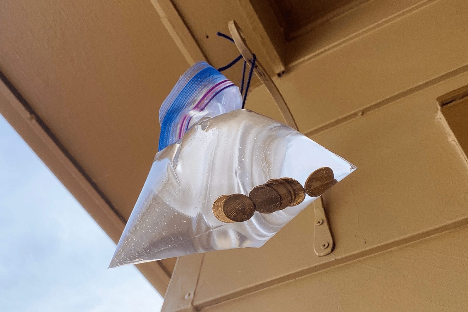 A plastic bag filled with water and coins is suspended from a structure, reflecting light in a partially cloudy outdoor setting.