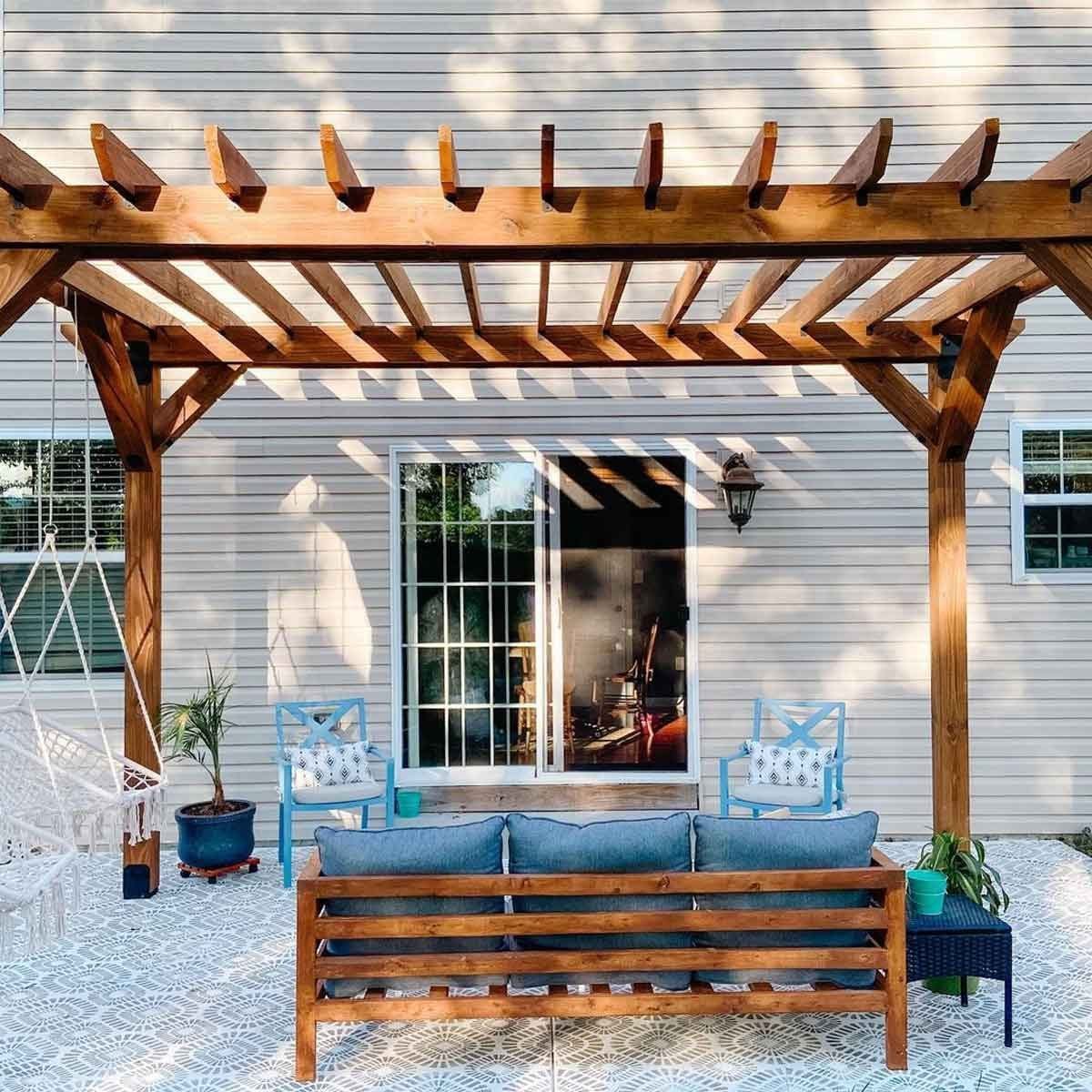 A wooden pergola shades a blue couch on a patterned patio, with a swing and chairs nearby, creating a cozy outdoor seating area beside a hou