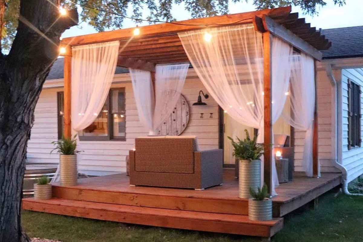 A wooden deck features a cozy seating area, draped in sheer fabric, illuminated by string lights, alongside potted plants and a welcoming ho