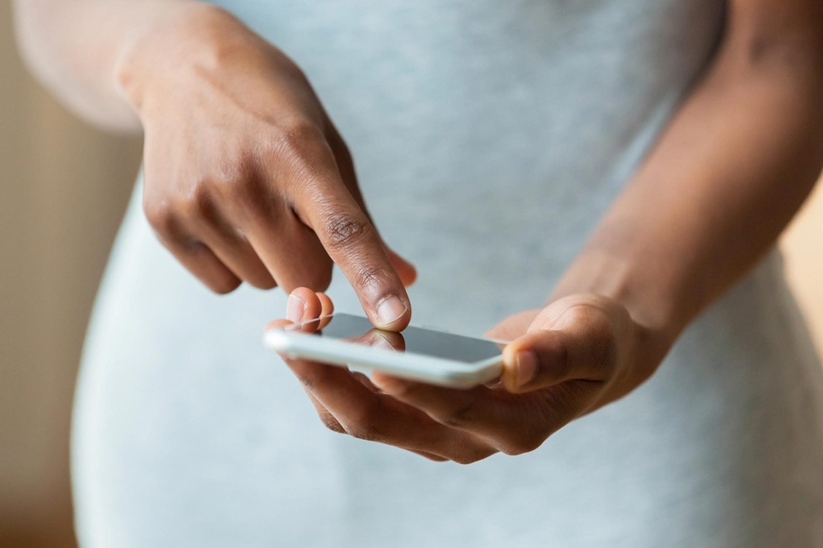 A hand holds a smartphone, with a finger touching the screen. The background is softly blurred, suggesting an indoor setting.