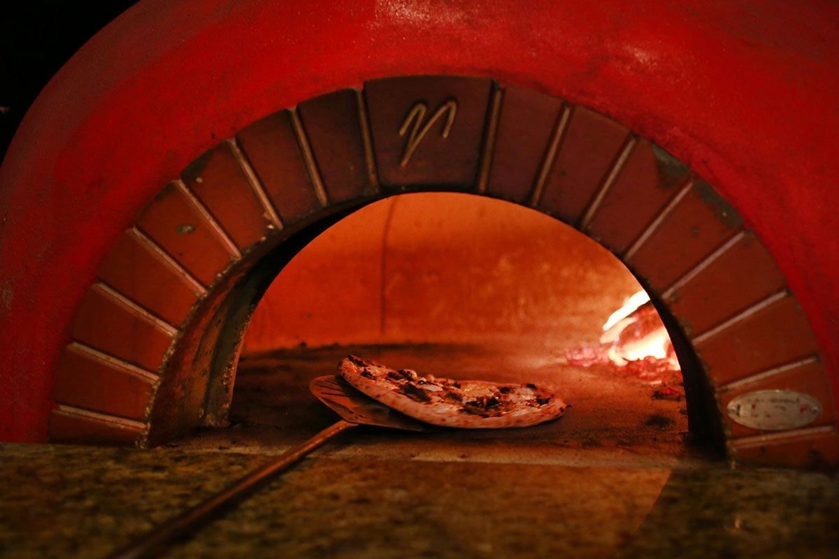 A pizza sits on a metal peel in front of a wood-fired oven, glowing with warm orange light. The oven’s red arch frames the scene.