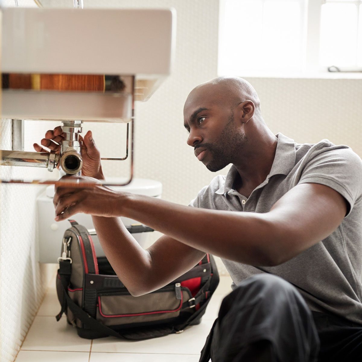 A man adjusts plumbing under a sink, focused on the task. A bag sits nearby in a bathroom with light-colored tiles and a white toilet.