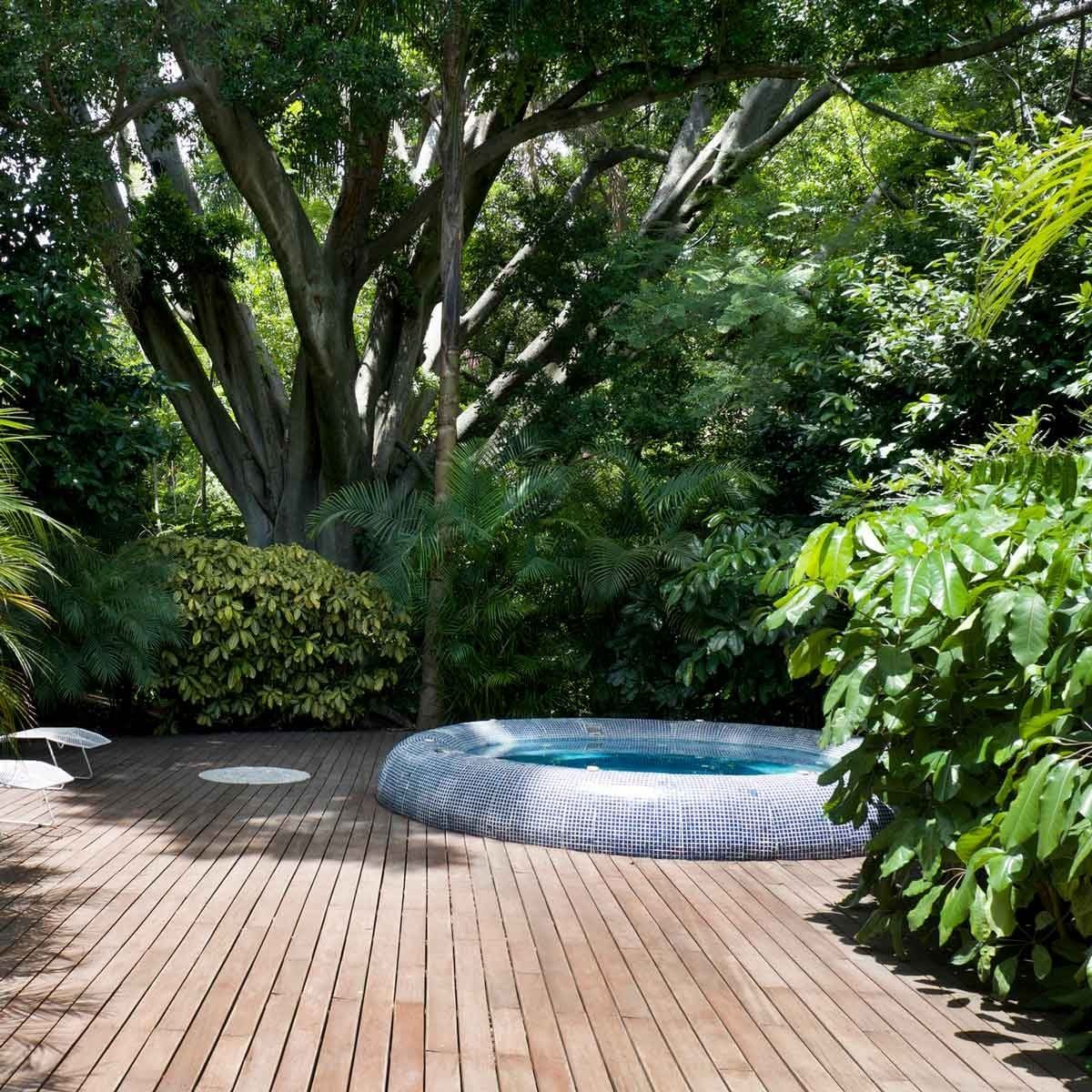 A circular jacuzzi sits on a wooden deck surrounded by lush greenery and large trees, creating a serene, secluded outdoor space.