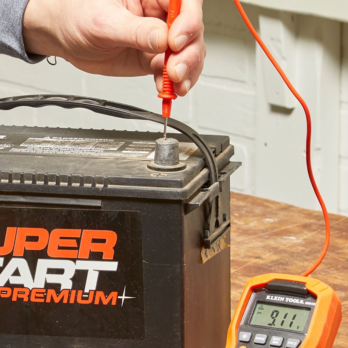 A hand holds a multimeter probe, testing a battery terminal while a digital multimeter displays a voltage reading on a wooden surface beneath it.