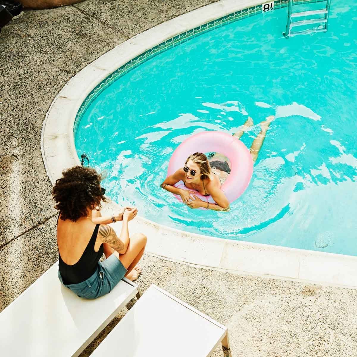 A woman relaxes in a pink float in a swimming pool, while another woman sits on a lounge chair nearby, enjoying the sun.