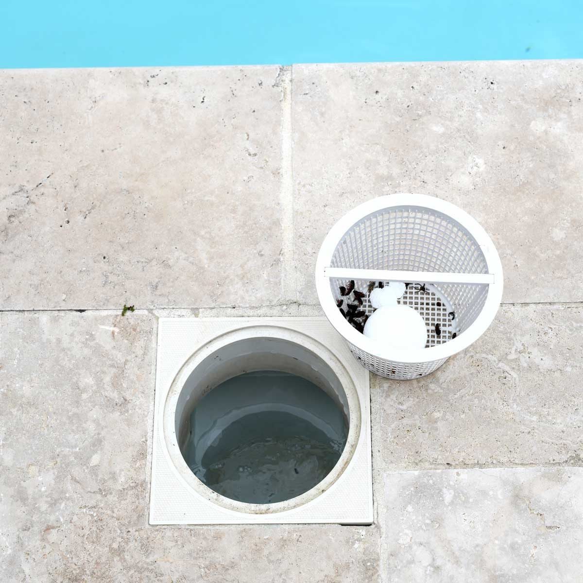 A circular drain is filled with water, adjacent to a white mesh waste basket containing debris, set on a stone surface beside a blue pool.