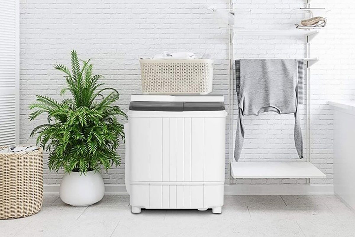 A white washing machine sits against a white brick wall, with a plant nearby and laundry hung on a shelf, situated in a clean, bright room.
