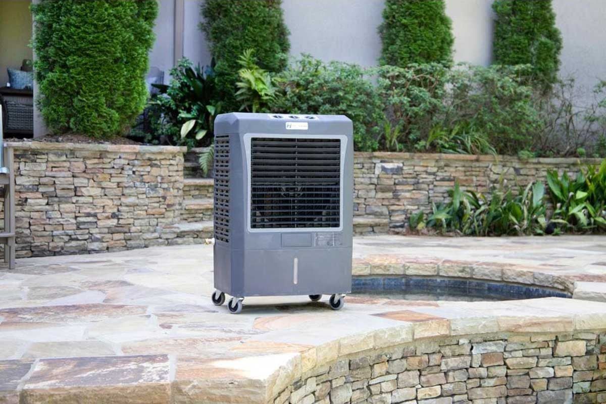 A gray air cooler stands on a stone patio, actively providing cool air in a well-kept outdoor space surrounded by lush greenery.