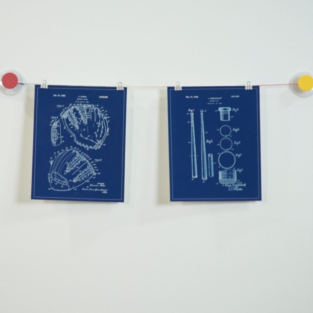 Two blueprints hang on a line, depicting designs for a baseball glove and other equipment, against a plain white wall.