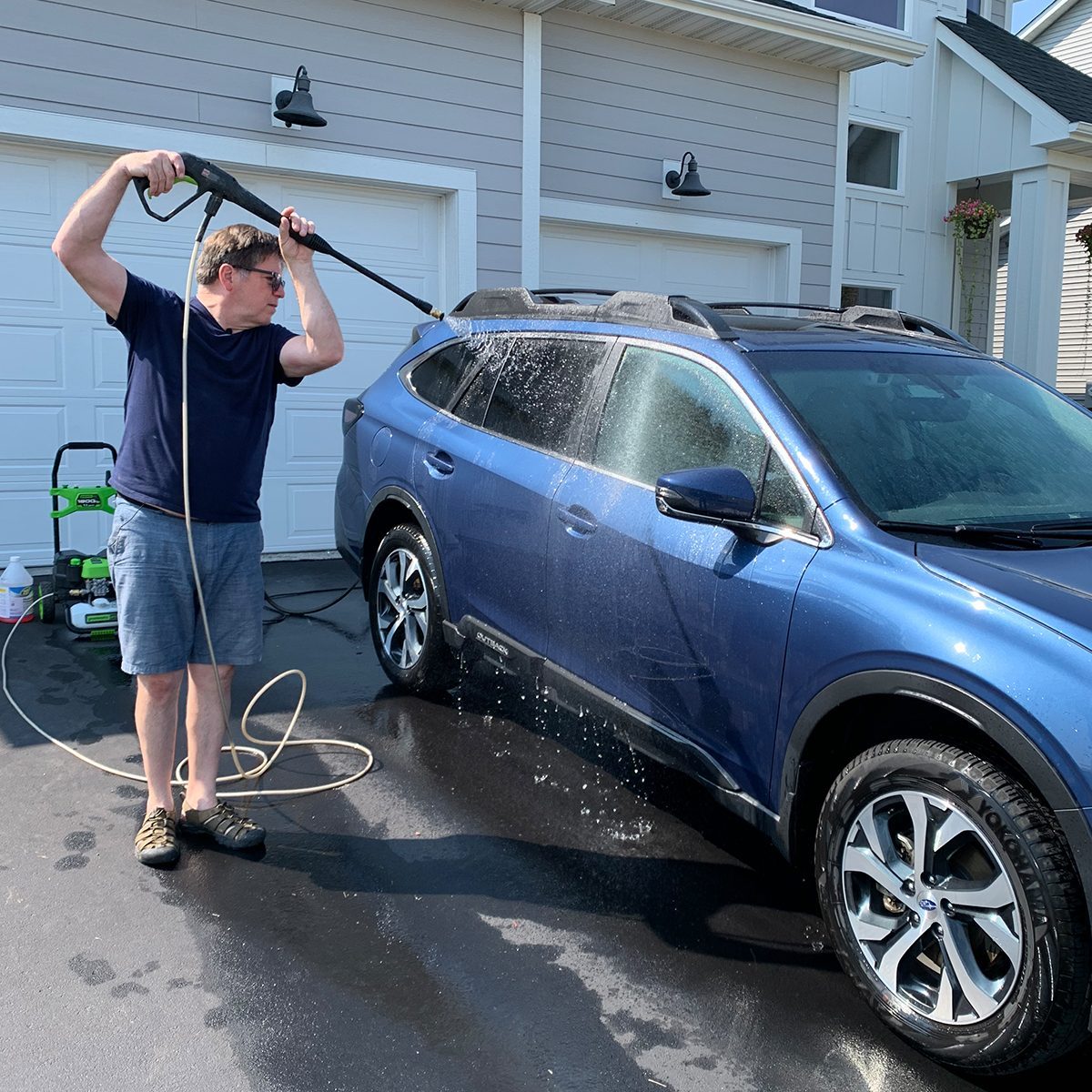 A man washes a blue car using a pressure washer outside a garage, with water spraying and driveway reflecting sunlight.