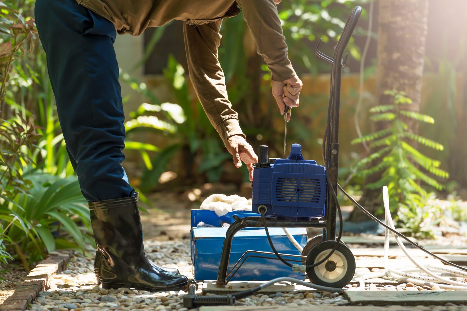 A person repairs a blue pressure washer while standing on gravel, surrounded by plants in a garden setting, wearing black rubber boots.