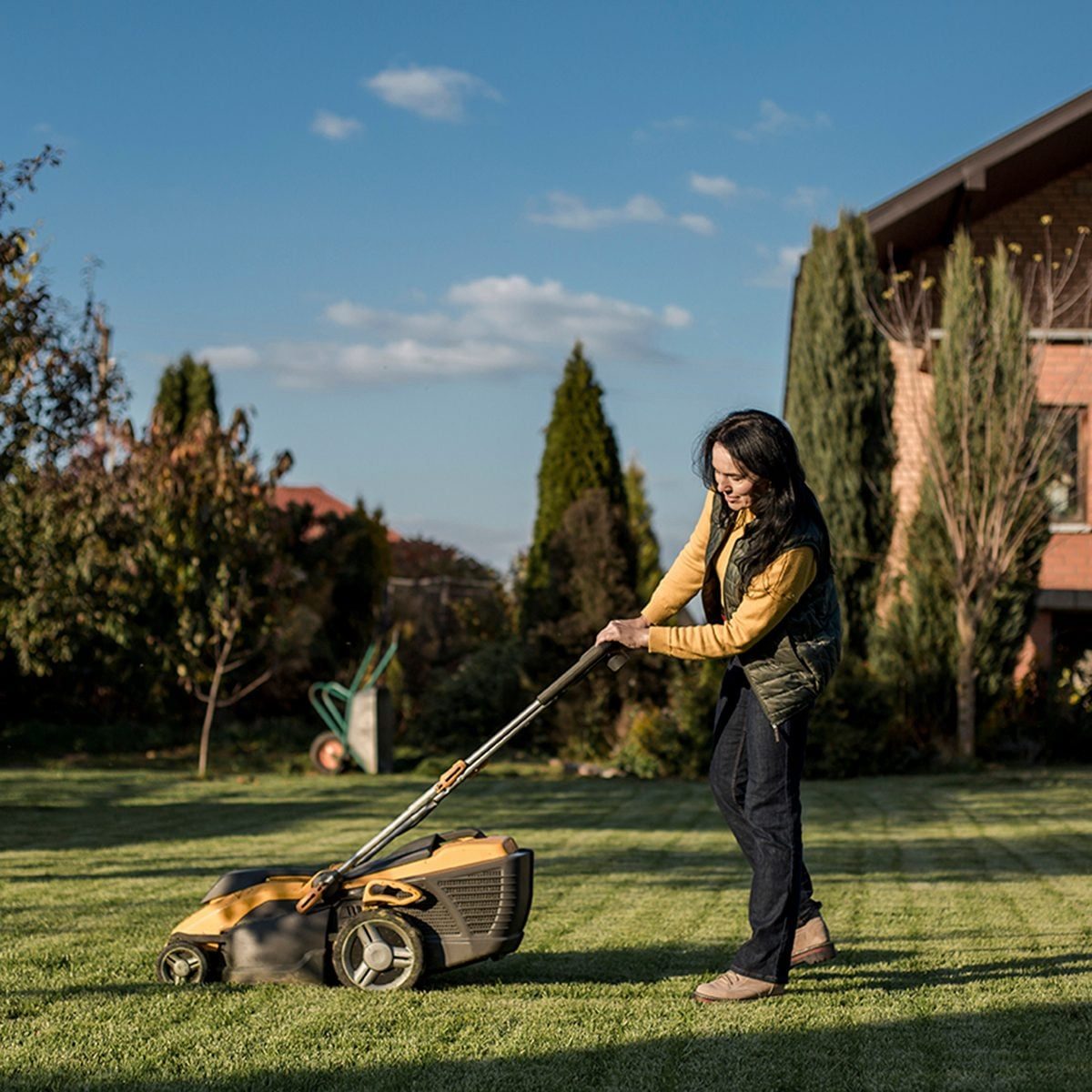 A woman mows the lawn with a yellow and black mower in a well-kept garden, surrounded by shrubs and a house under a clear blue sky.