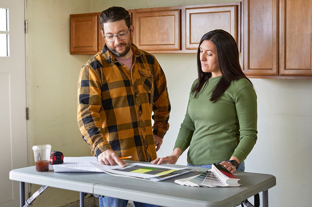 A man and woman stand at a table looking at papers and color samples, appearing to discuss home renovation plans.