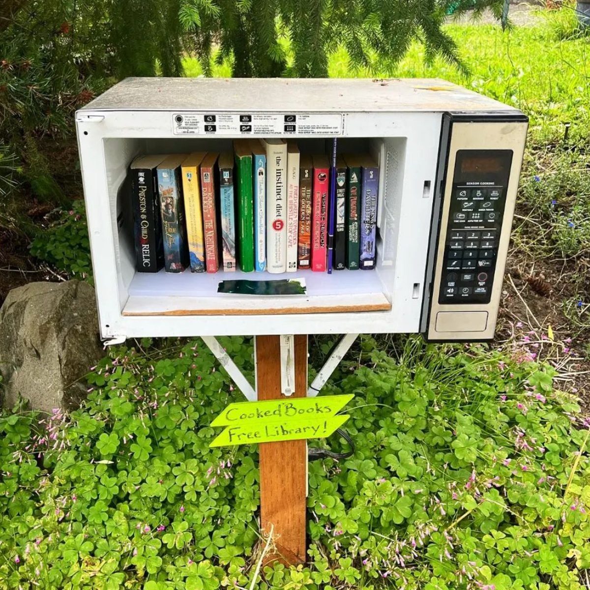 A repurposed microwave contains various books inside, displayed outdoors amidst greenery, with a sign stating "CookedBooks Free Library!" ne