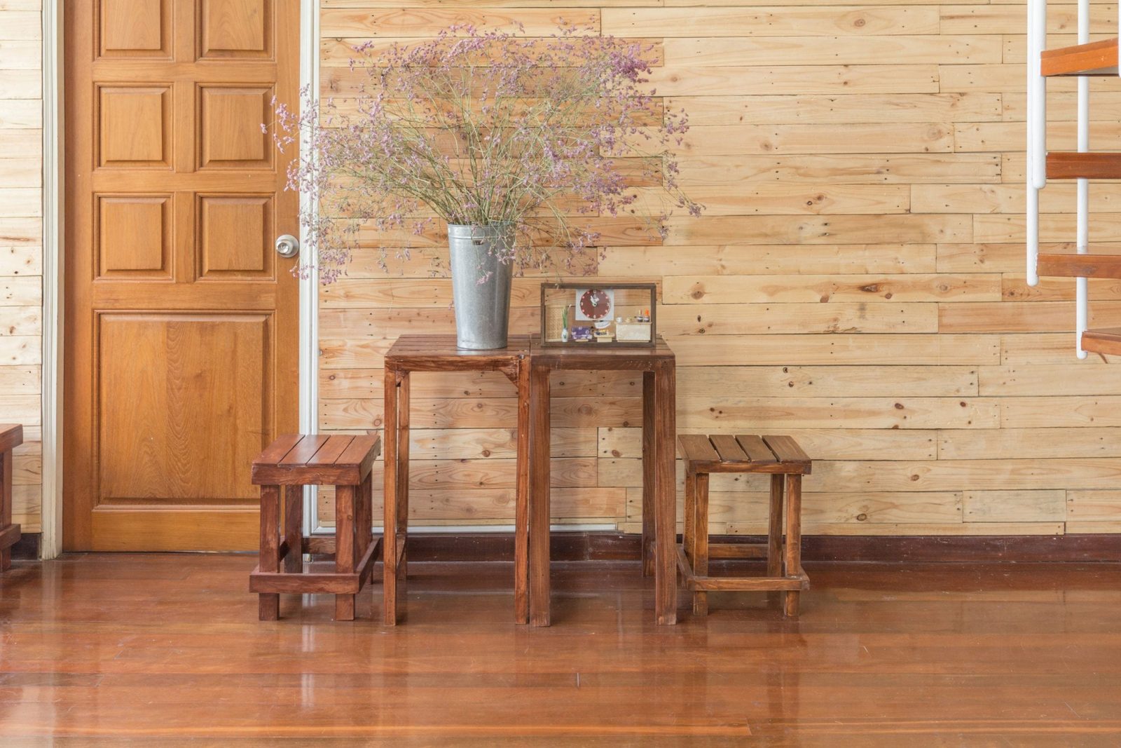 A wooden table holds a vase of dried flowers and a framed photo, surrounded by two matching stools, against a light wood-paneled wall and do