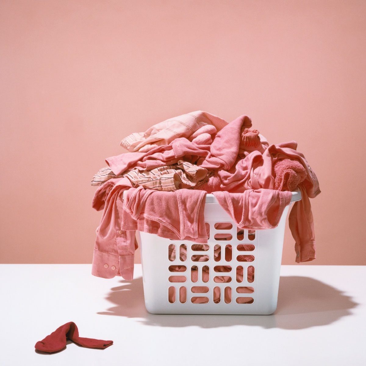 A white laundry basket overflows with pink and red clothing on a simple table, set against a soft pink background, evoking a cozy, domestic