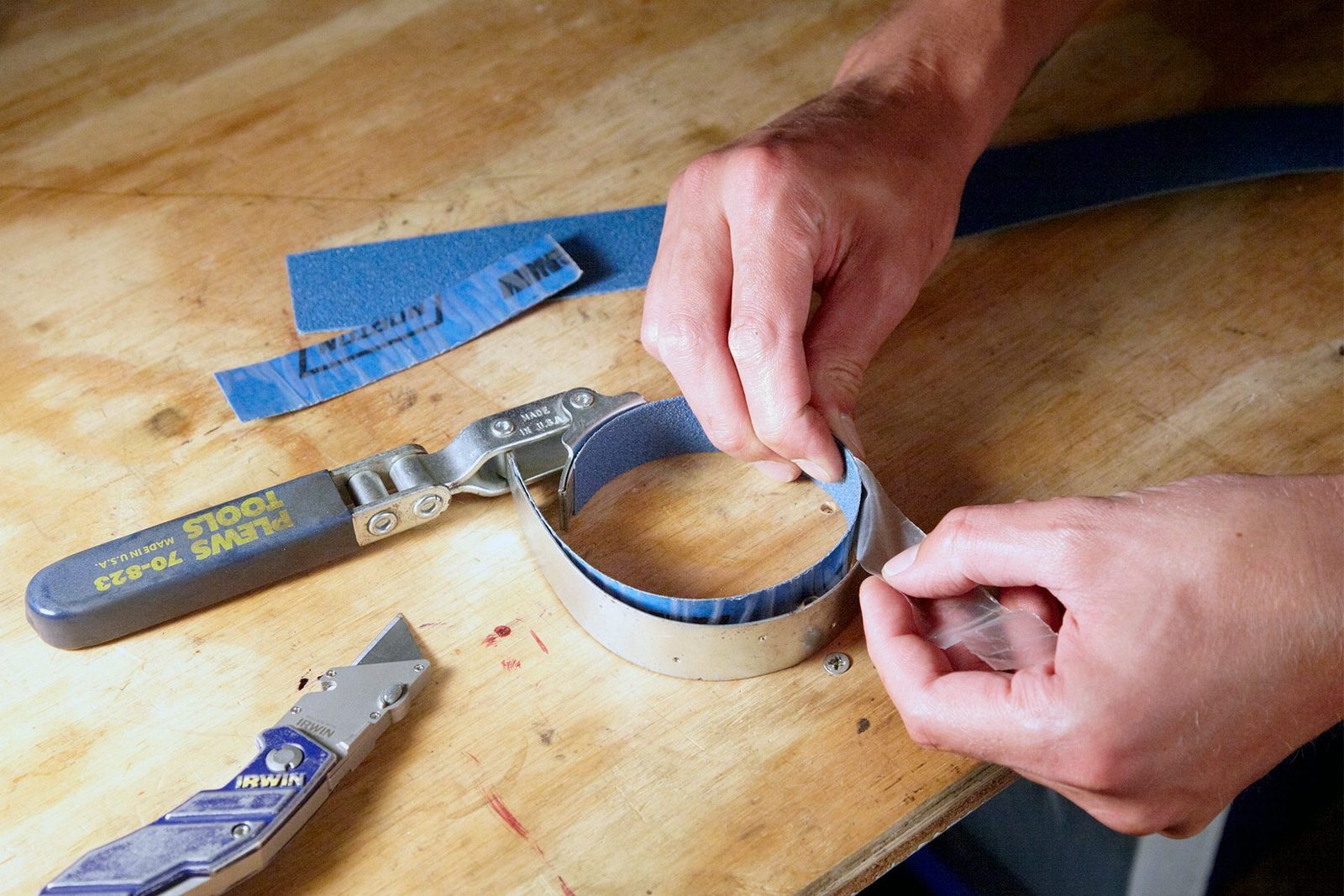 A close-up image of hands working on a wooden surface with tools. One hand is gripping a piece of blue sandpaper, while the other peels back a layer from it. A utility knife and