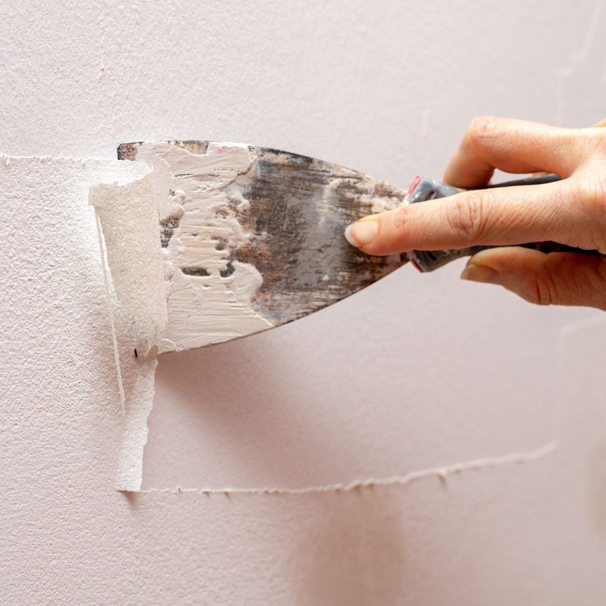 A hand holds a putty knife, applying a layer of spackle to a textured, light-colored wall. The action smooths the surface for finishing.