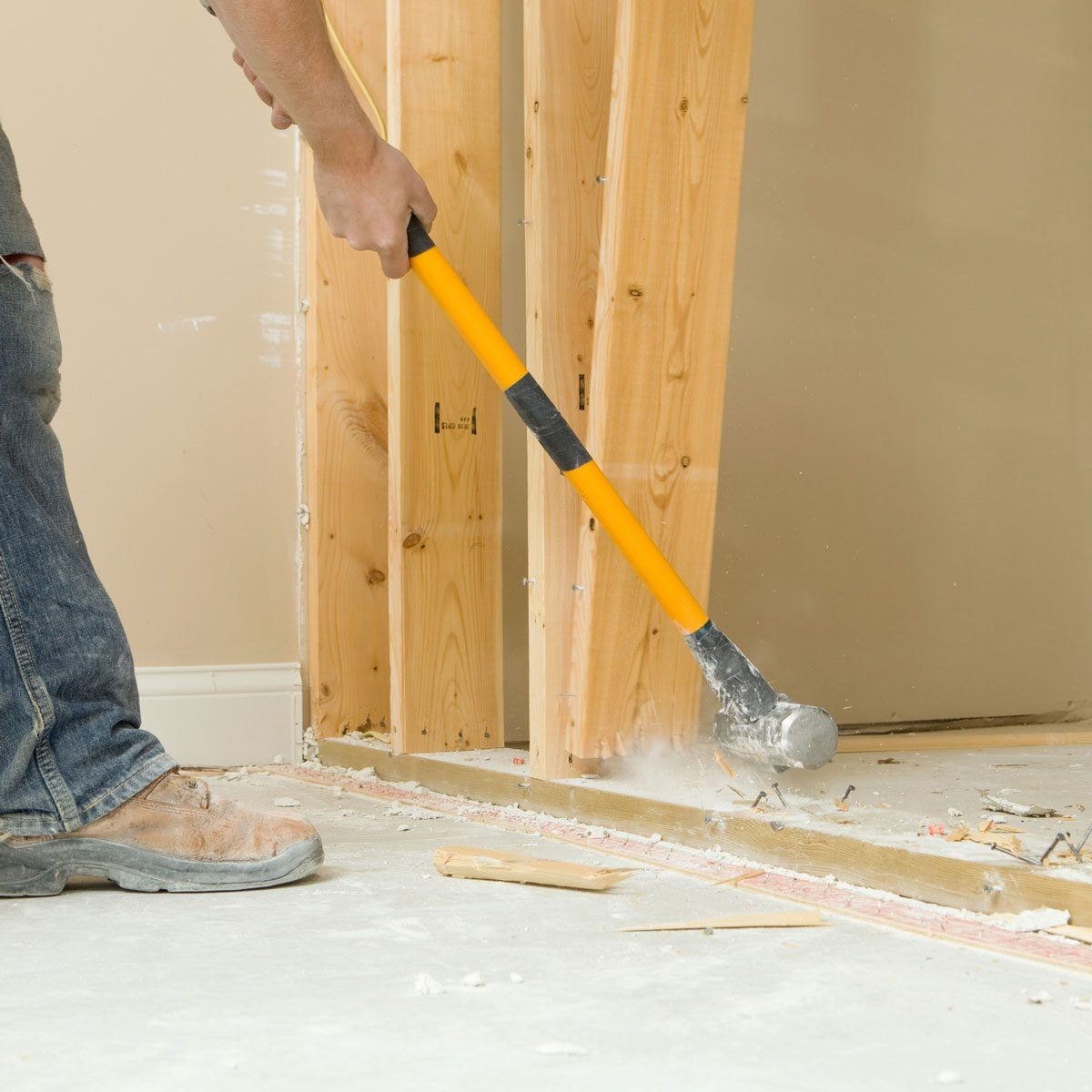 A person swings a demolition hammer, breaking apart flooring in a construction area surrounded by wooden studs and debris on a light-colored