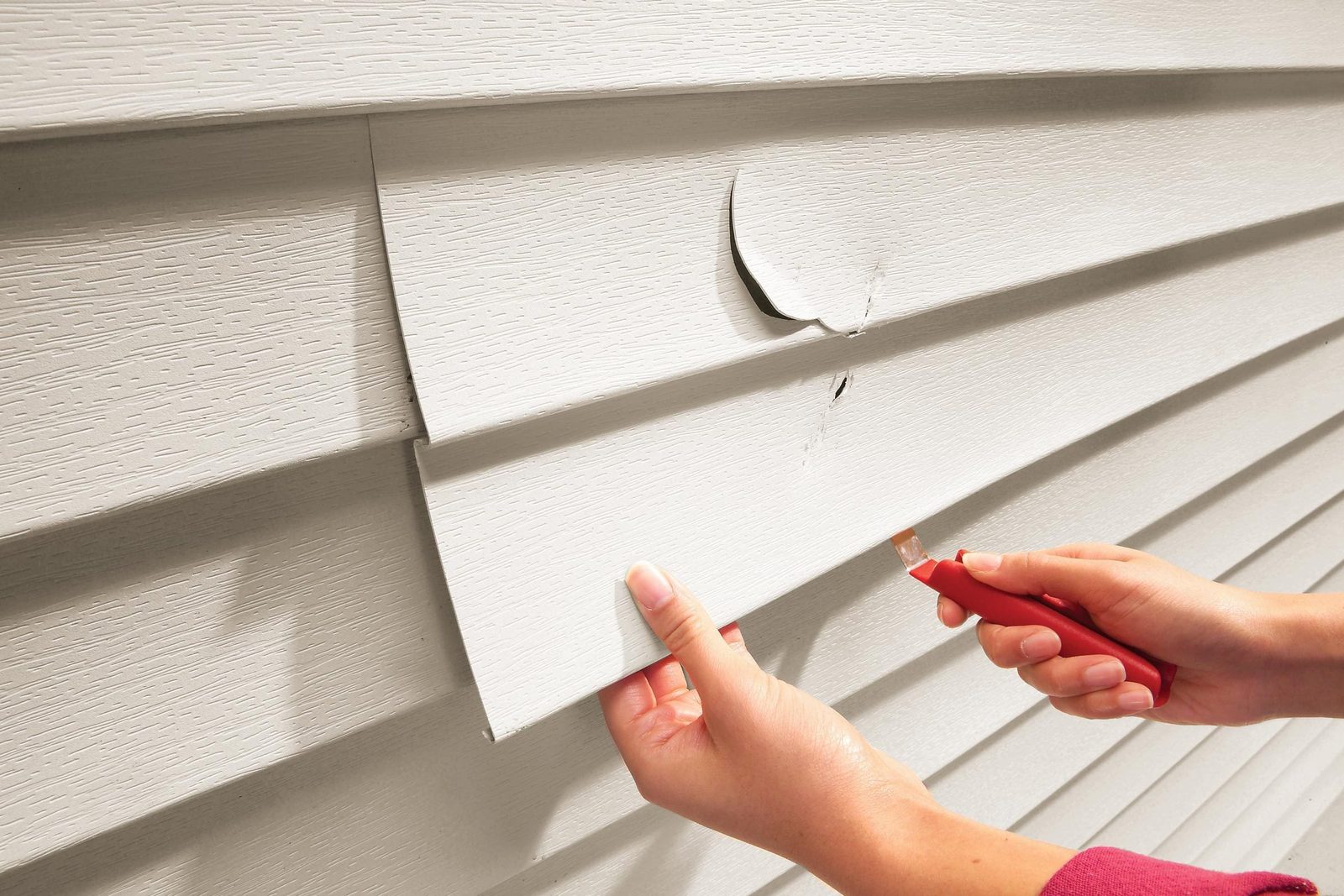 A hand holds a red tool, removing a vinyl siding piece from a wall with visible damage, showing the textured surface and shadows.
