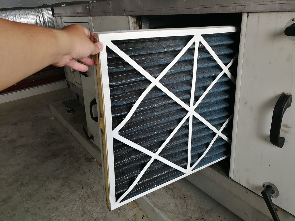 A hand holds a rectangular air filter, removing it from a machine in a clean, industrial setting with concrete flooring and metal equipment