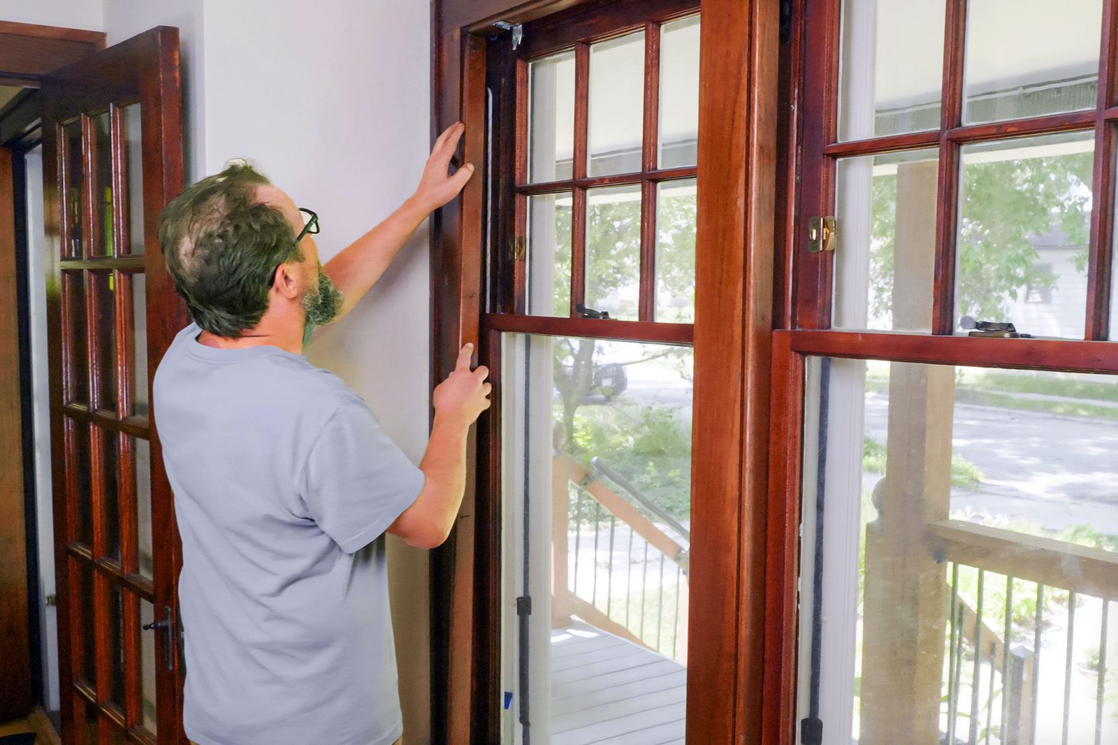 A man adjusts the frame of a wooden window, indoors, while sunlight filters through the glass, illuminating the surrounding room and visible