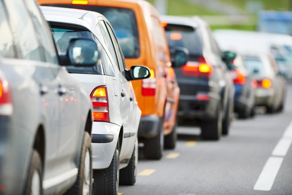 Cars are stopped in traffic, facing forward on a multi-lane road, surrounded by additional vehicles in the background and greenery on the si