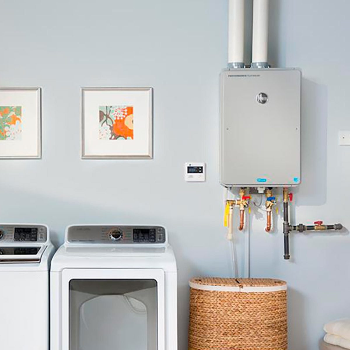A washing machine and dryer sit side-by-side against a light blue wall, accompanied by framed artwork, a water heater, and a woven laundry b
