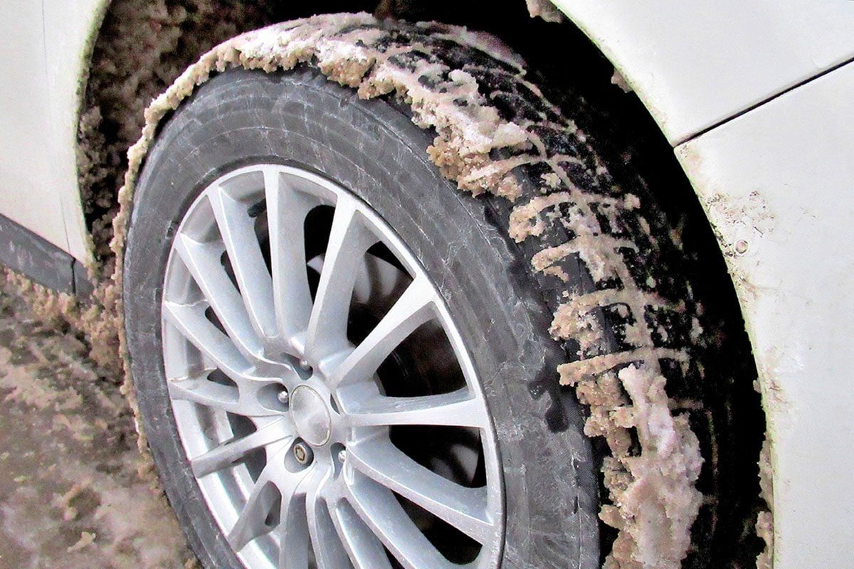 A dirty tire, covered in salt and grime, is positioned next to a white vehicle, indicating usage in snowy or icy conditions.