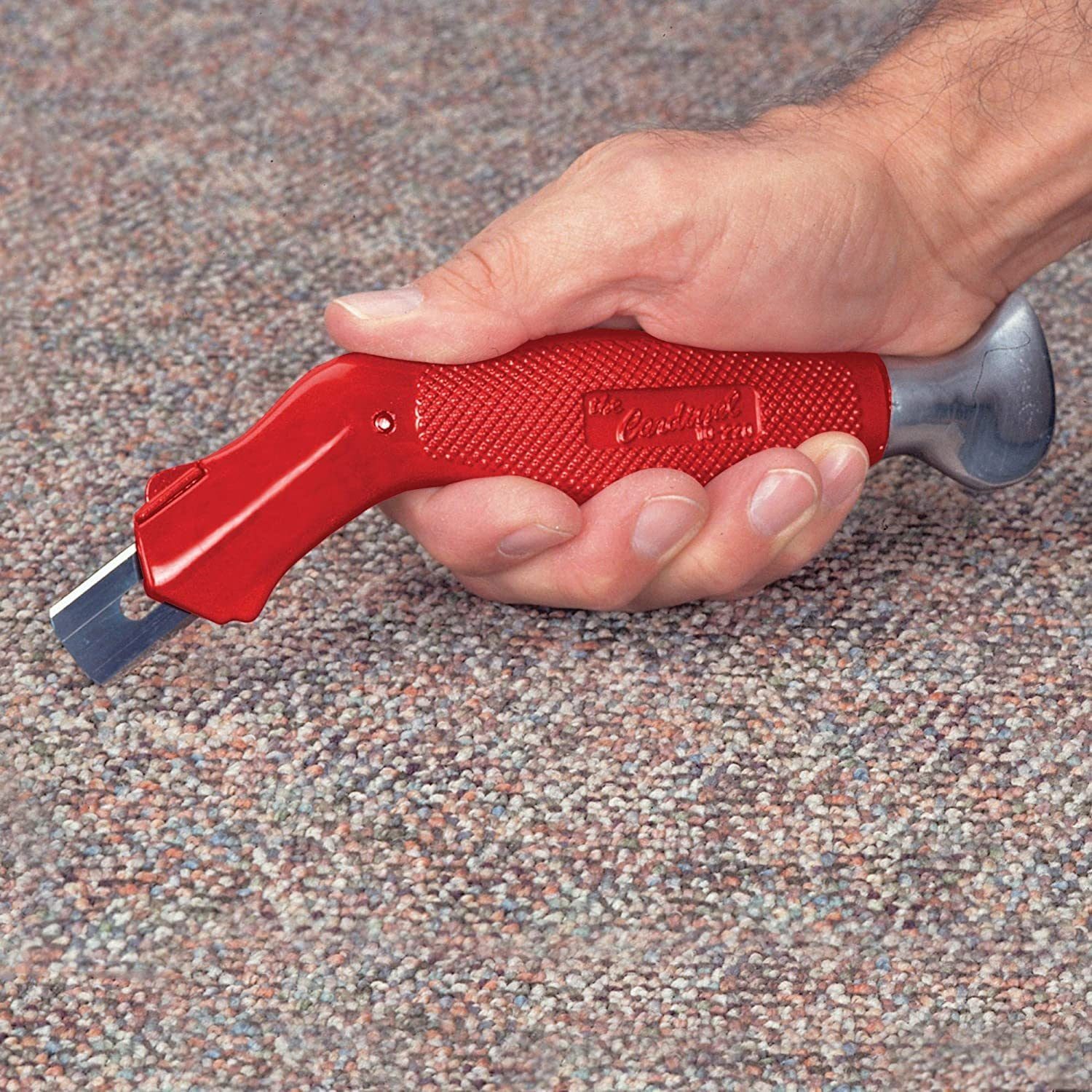 A red carpet knife is held above a textured carpet surface, positioned to cut through the material while being gripped securely by a person'