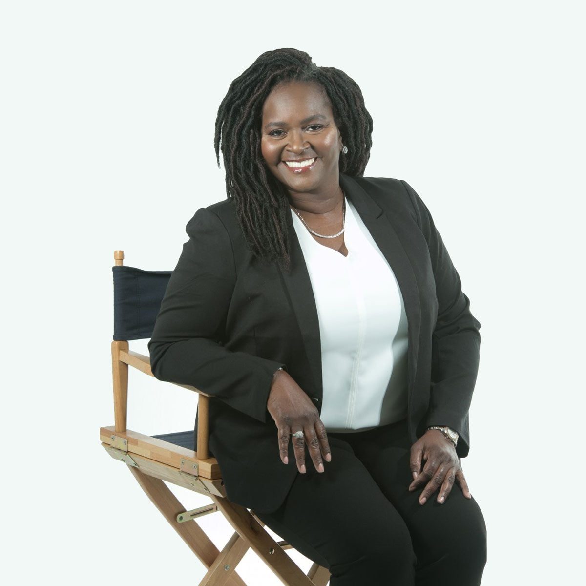 A woman in a black suit sits confidently on a director's chair, smiling against a light, neutral background.