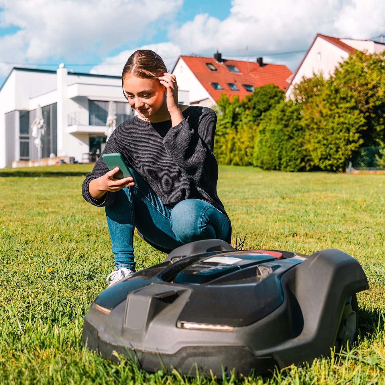 A robotic lawn mower sits on green grass. A young woman crouches nearby, smiling and using her smartphone in a sunny, residential yard.