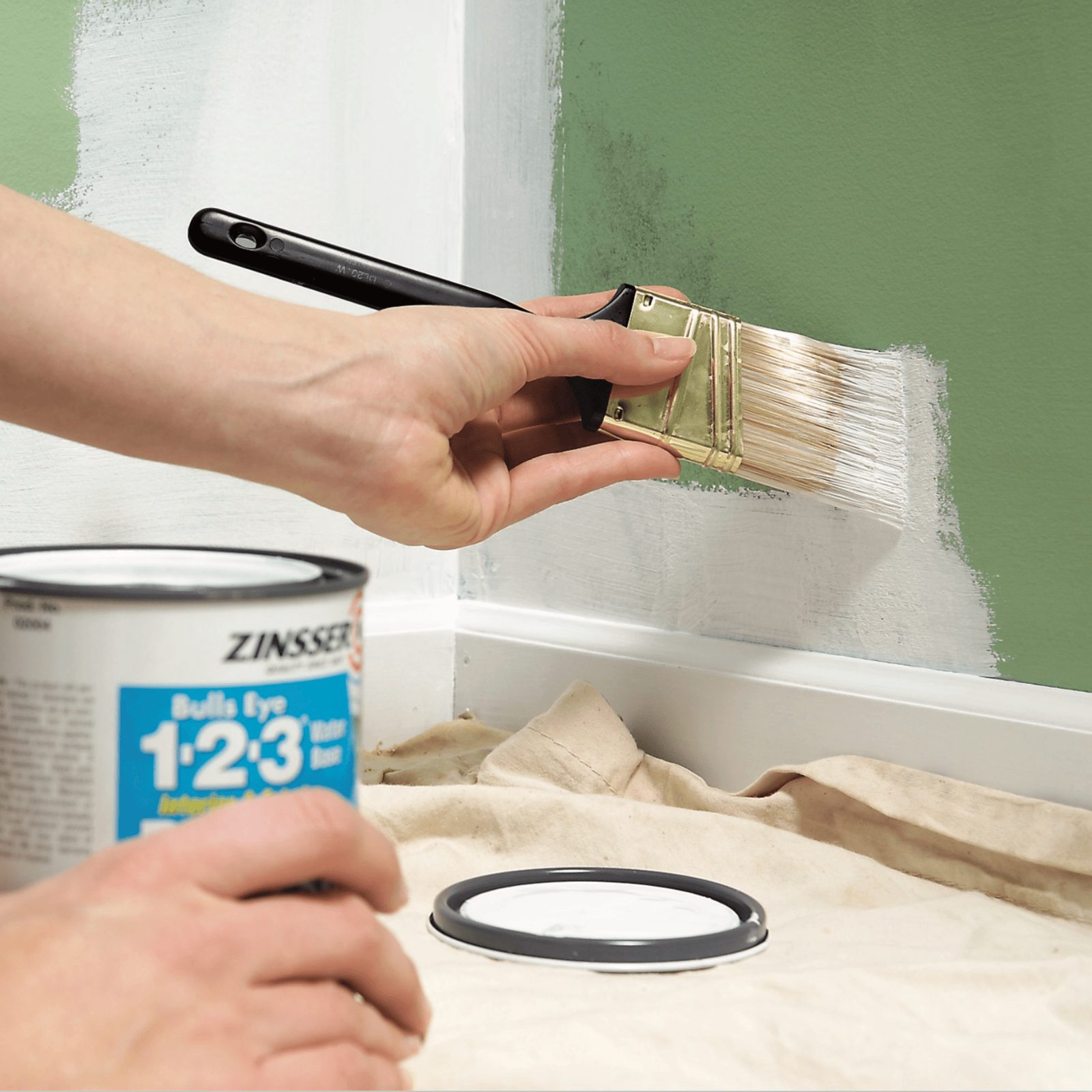A hand paints a wall with a brush, while holding a paint can nearby. The area is covered with a drop cloth.