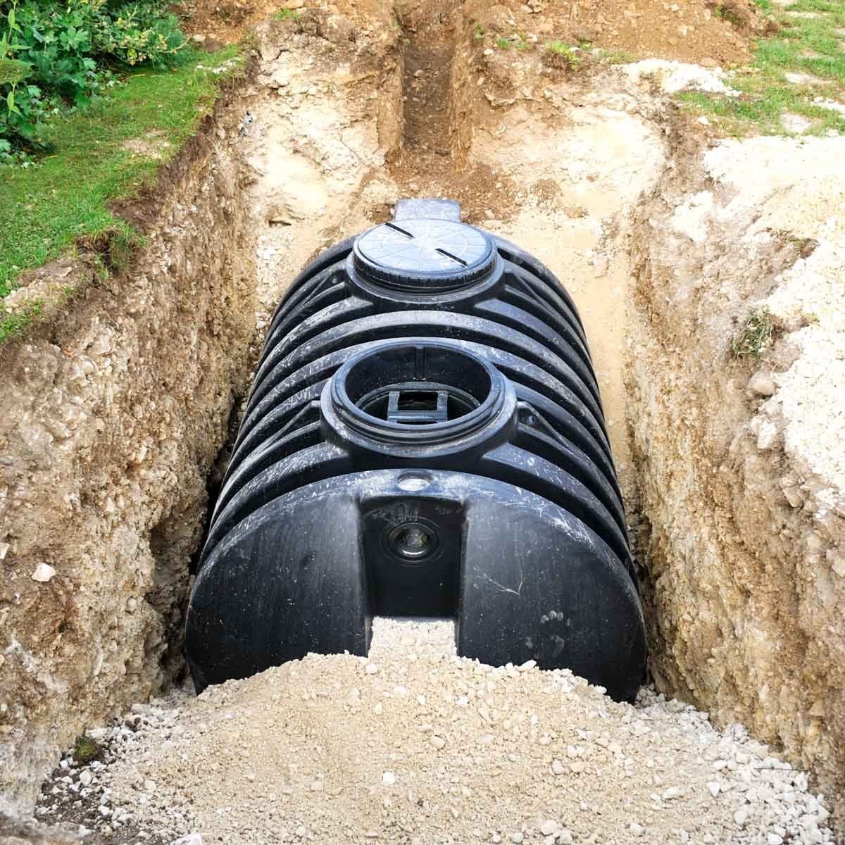 A large black tank lies in a freshly dug trench, partially covered with dirt, surrounded by grass and gravel, indicating installation for dr