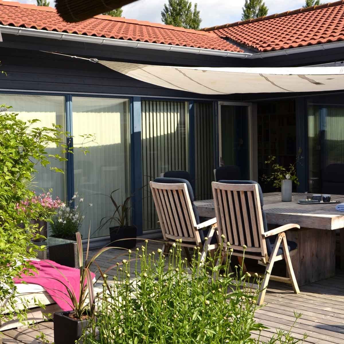 A wooden table with chairs sits on a wooden deck, surrounded by greenery and potted plants, under a sunshade in a bright outdoor setting.