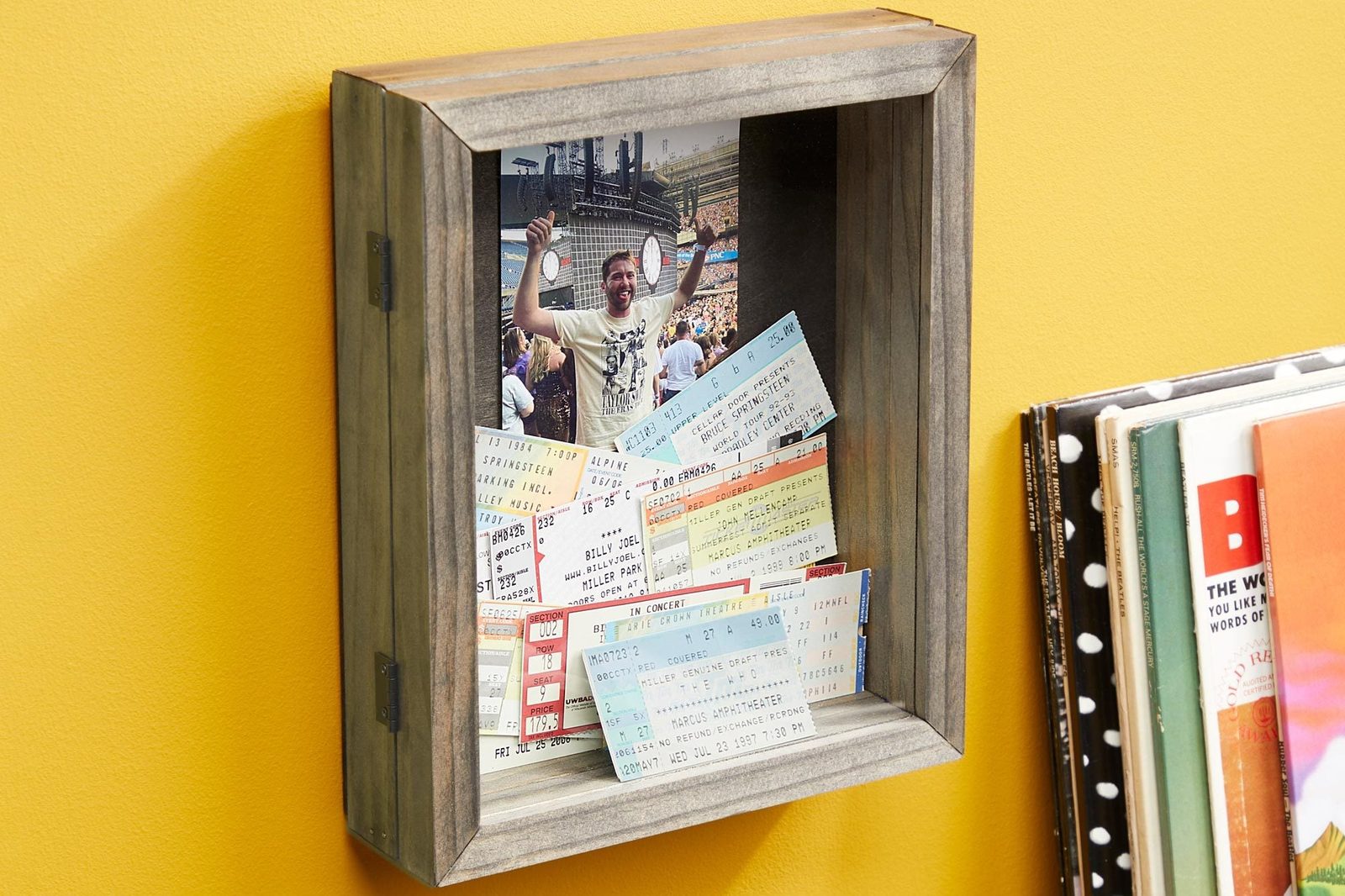 A wooden shadow box displays a concert photo and various event tickets, set against a bright yellow wall, conveying vibrant memories of musi