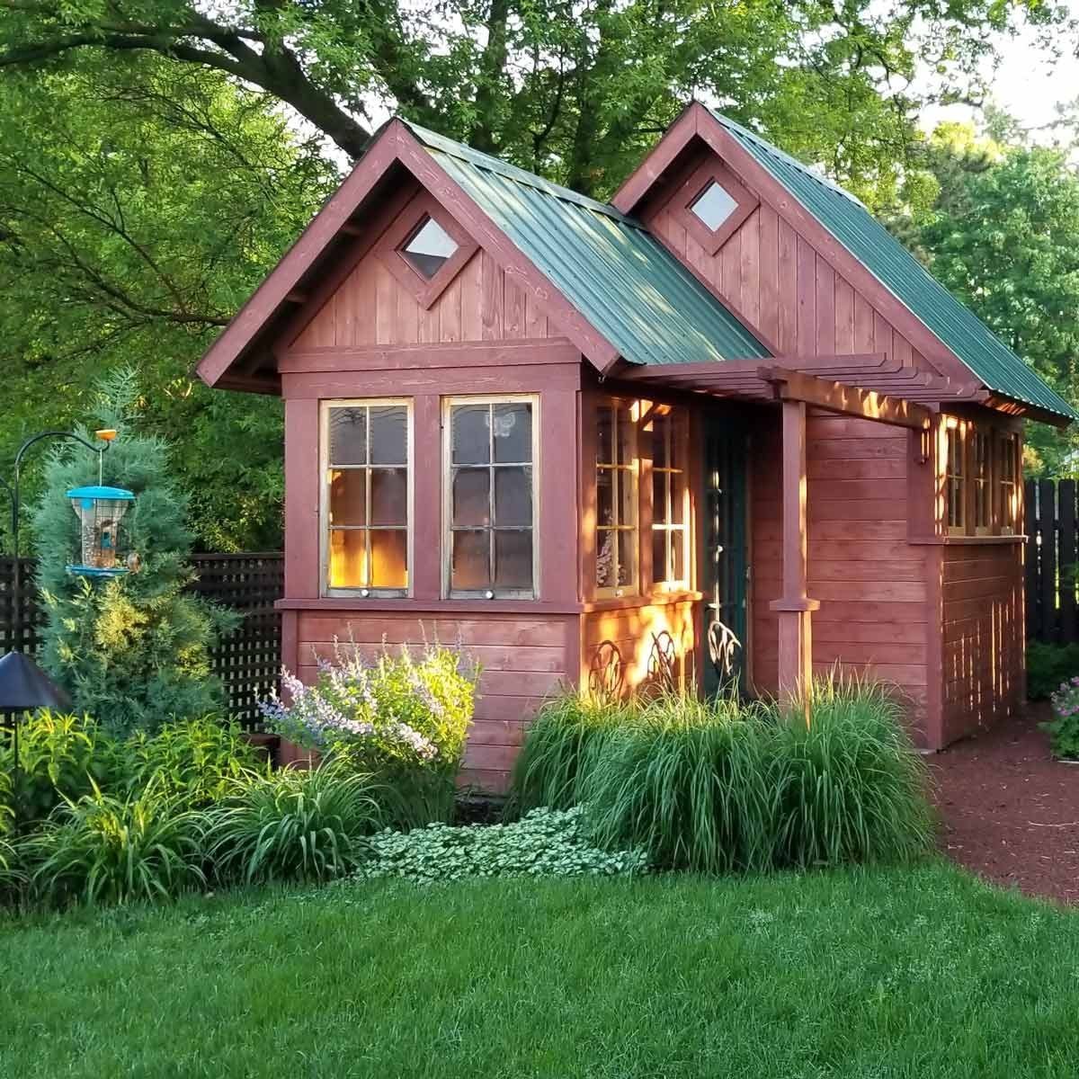 A charming, wooden shed with a green metal roof stands amidst lush grass and flowering plants, illuminated by sunlight in a serene garden se