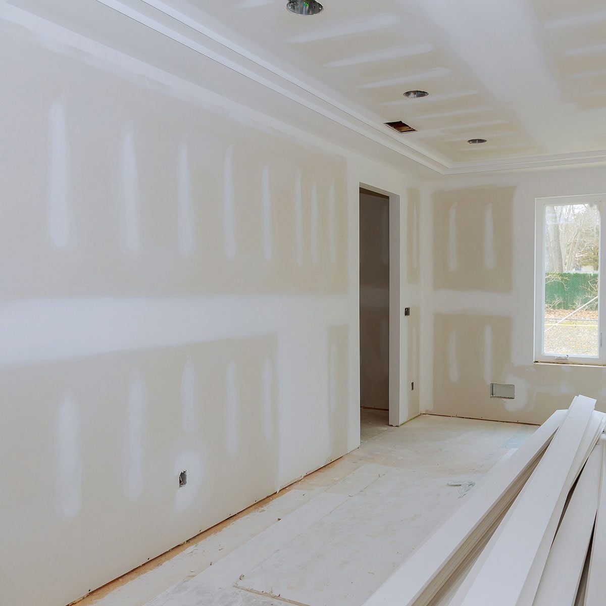 A room under renovation showcases unfinished drywall on the walls, with white boards stacked on the floor and a window revealing an outdoor